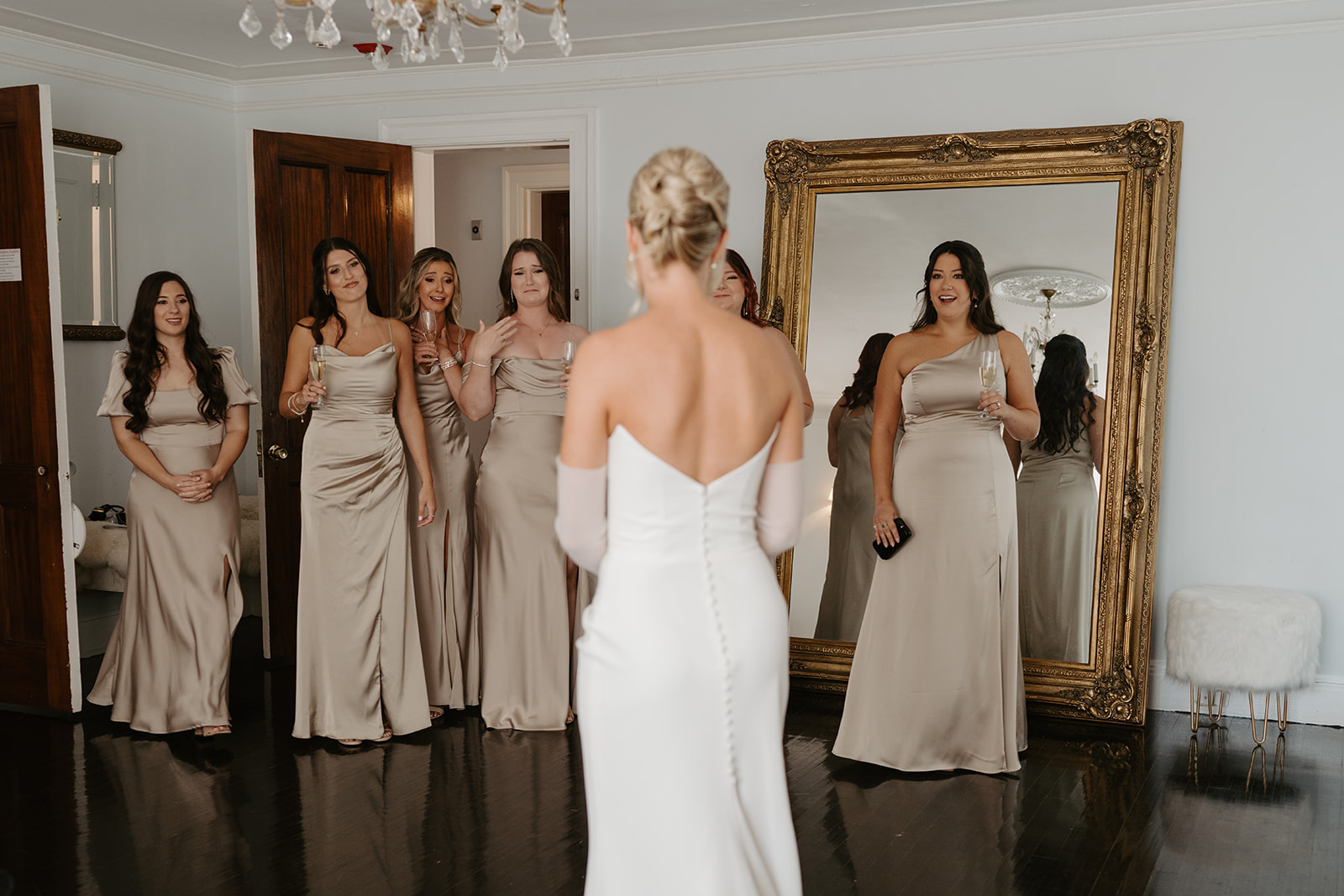Bridesmaids in champagne dresses react with emotional smiles to the bride's first look in her gown.